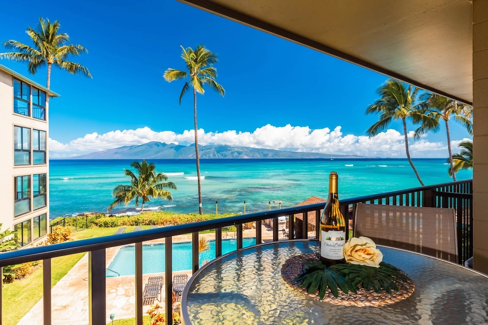 A vacation rental balcony overlooking a resort pool with ocean and mountain views in the background